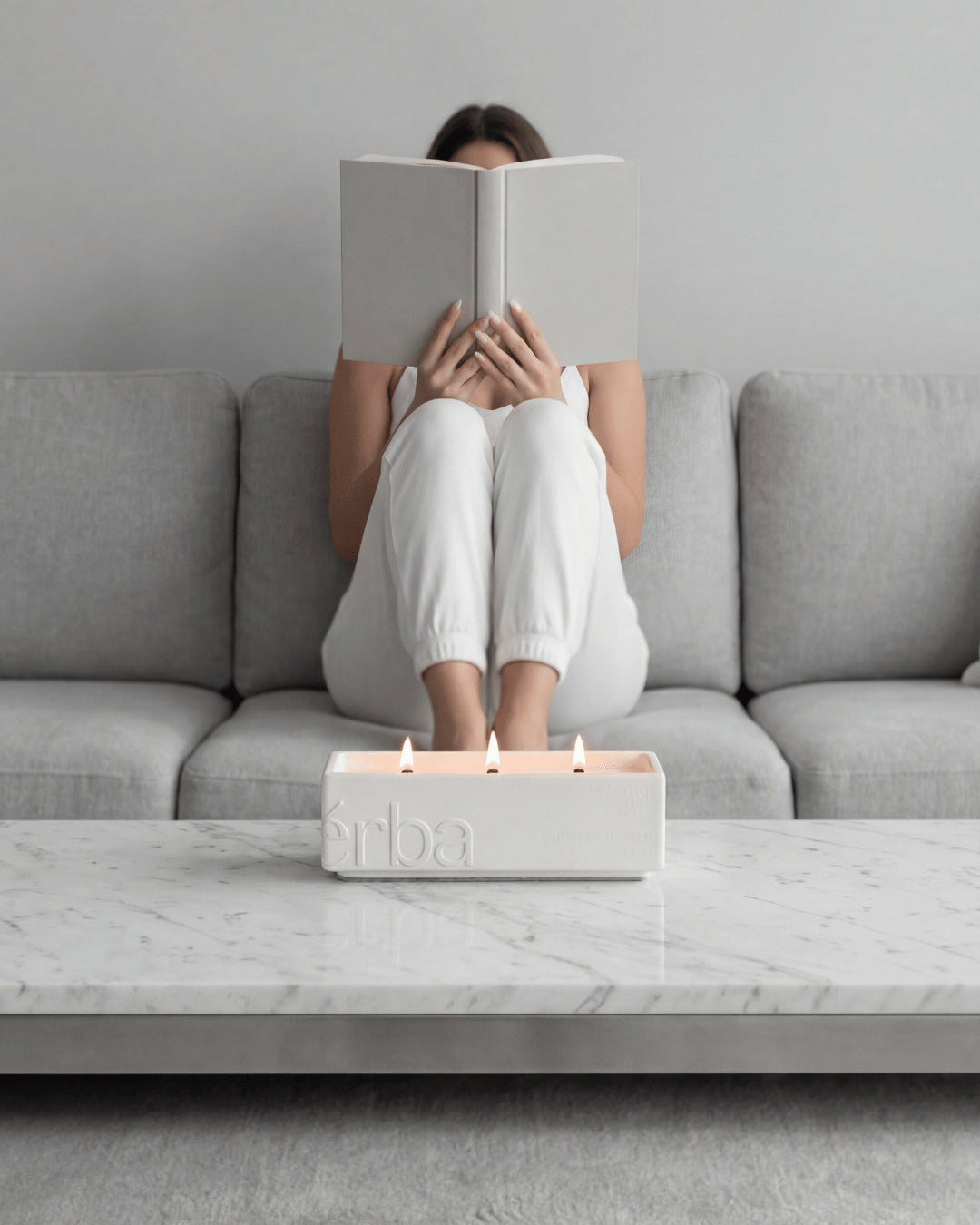 Person reading a book on a grey sofa behind a large white Vérba three-wick candle on a marble table