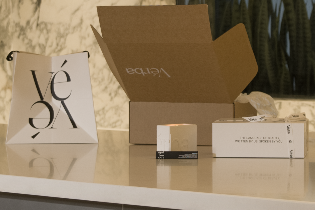 Verba packaging setup featuring a branded gift bag, shipping box, and candle on a marble surface — luxury unboxing scene.