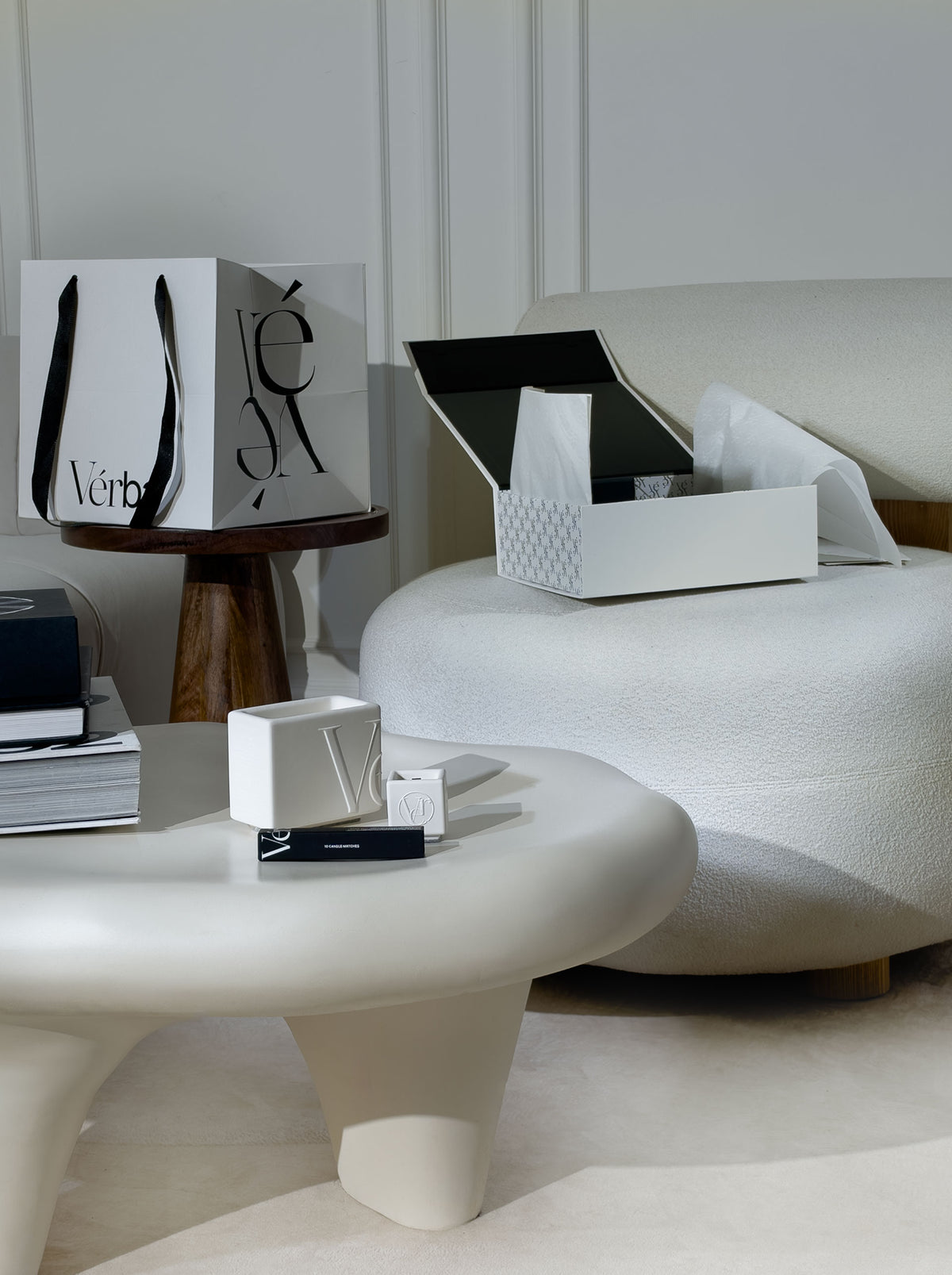 Vérba White Collection candles on a sculptural white coffee table, with a white gift box and shopping bag in a minimalist interior