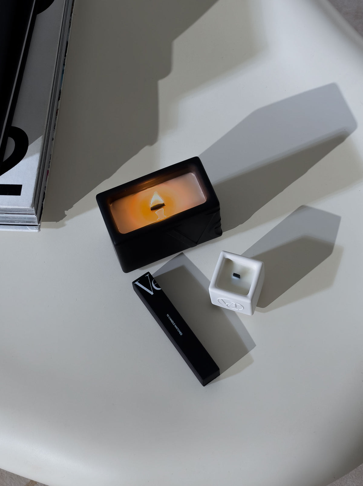 Lit Verba black candle with wooden wick, black matchbox and white mini tester candle on a cream table — top view
